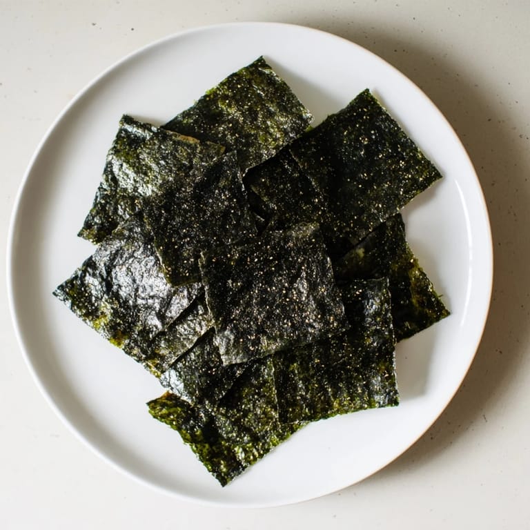 Close-up of crispy roasted seaweed chips with dark green color, scattered salt flecks and a pinch of chili flakes for heat.